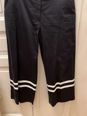 Marinblå vida byxor med vita ränder - Säljer ett par marinblå vida byxor med två vita ränder längst ner på benen. Byxorna har hög midja och raka ben, samt sidofickor. Materialet är mjukt och lite glansigt, vilket ger en sportig och trendig look.