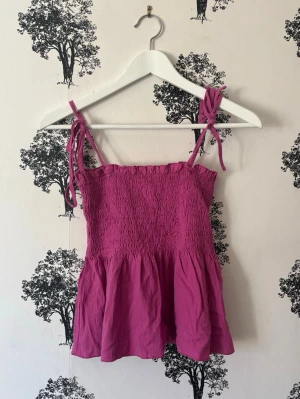 Rosa linnetopp med knytband - Supersöt rosa linnetopp med smockad överdel och volang nertill. Axelbanden knyts på axlarna för en extra gullig touch. Perfekt för varma dagar och enkel att styla med jeans eller kjol.