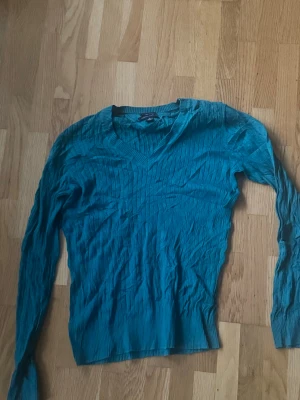 Turkos stickad tröja med V-ringning - Snygg turkos stickad tröja med V-ringning och ribbade muddar. Tröjan har lång ärm och ett diskret mönster i stickningen som ger en cool struktur. Perfekt att matcha med jeans eller kjol för en chill look.