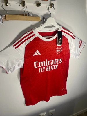 Arsenal matchtröja  Adidas - Snygg Arsenal matchtröja från Adidas i rött och vitt med klubbmärke och sponsortryck på bröstet. Tröjan har vita ärmar, röda axelränder och är tillverkad i lätt funktionsmaterial. Perfekt för fotboll eller träning.