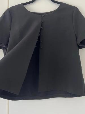 Svart blus med knappar framtill  - En svart blus med rund halsringning och korta ärmar. Framsidan har en rad klädda knappar som ger en stilren och modern känsla. Blusen har en lätt A-linjeformad passform och är perfekt för dig som gillar minimalistisk stil. Så fin till våren och sommaren! 
