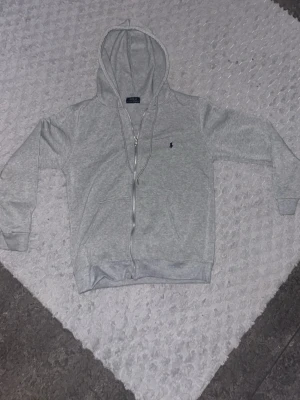 Grå zip-upp från Polo Ralph Lauren - Ljusgrå zip-upp med dragkedja från Polo Ralph Lauren. Klassisk design med huva, fickor framtill och blå broderad logga på bröstet. Tillverkad i mjukt bomullsmaterial som är skönt att ha på sig. Perfekt för en avslappnad och stilren look. Knappast använd!!!