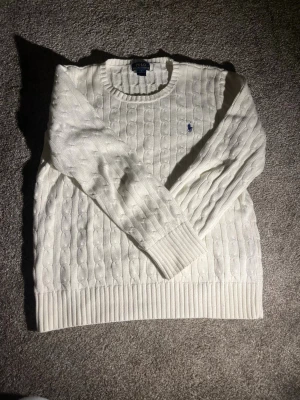 Vit kabelstickad tröja Polo Ralph Lauren - Vit kabelstickad tröja från Polo Ralph Lauren med rund hals och blå broderad logga på bröstet. Tröjan har långa ärmar och breda ribbade muddar vid ärmslut och nederkant. Klassisk och tidlös design i mjukt material. Tröjan är storlek junior l 
