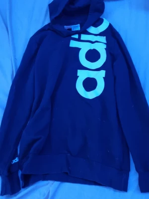 Svart hoodie från Adidas med tryck - Snygg svart hoodie från Adidas med stort vitt logotyptryck längs framsidan. Klassisk modell med huva och ribbade muddar. Perfekt för en sportig och avslappnad stil. Materialet är mjukt och bekvämt, passar till både träning och chill.