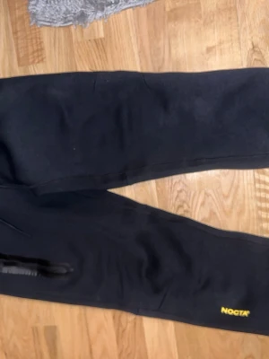 Svarta mjukisbyxor från NOCTA - Säljer ett par svarta mjukisbyxor från NOCTA med gul logga på benet. Byxorna har en avslappnad passform och dragkedja vid fickan. Perfekta för chill dagar och streetwear-stil. Pris kan diskuteras 