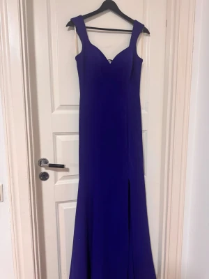 Lång blå klänning med slits - Elegant långklänning i djupblå färg med breda axelband och v-ringad urringning. Klänningen har en stilren design med en diskret slits framtill. Perfekt för speciella tillfällen när du vill sticka ut med en klassisk look.