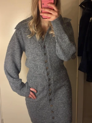 Grå stickad klänning från Adoore - Mysig grå stickad klänning från Adoore med markerad midja, stora krage och knappar framtill. Klänningen har lång ärm och är i mjukt material. Enkel men stilren design som är lätt att matcha.