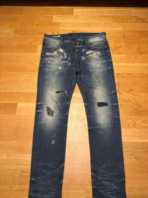 Blå  jeans från Scotch & Soda - Säljer ett par blå jeans från Scotch & Soda, modell Ralston. Jeansen har en slim fit-passform, slitna detaljer, hål och patchade partier samt ljusa tvättade partier framtill från fabrik. Klassisk femficksmodell med knappgylf och coola kontrastsömmar. Strl 30/32