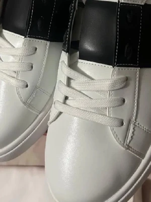 Vita sneakers med svart detalj 43 - Snygga vita sneakers med bred svart rem över snörningen och coola nitar på sulan. Skorna har klassisk låg profil och är tillverkade i slätt lädermaterial. Perfekta för dig som gillar stilrena och trendiga detaljer. skickar idag eller imorgon 😊