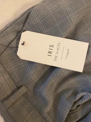 Grå rutiga kostymbyxor IRIS - Snygga grå kostymbyxor från Lindex, modell IRIS THE SLACKS. Byxorna har ett klassiskt rutmönster i vitt och är croppade med raka ben. Perfekta för en stilren look och har hällor för bälte samt fickor bak.