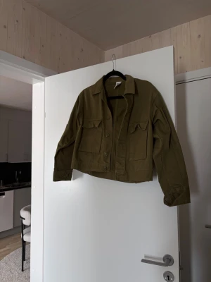 Olivgrön cropped overshirt - Snygg olivgrön overshirt i croppad modell med stora bröstfickor och knappar framtill. Tillverkad i ett kraftigare bomullstyg som ger en cool utility-vibe. Perfekt att slänga över en t-shirt för en avslappnad look.