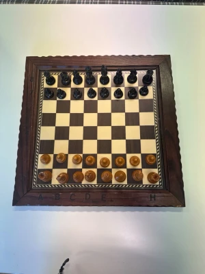 Schackspel i trä med bräde och pjäser - Säljer ett schackspel med schackbräde i trä och schackpjäser i svart och ljust trä. Brädet har bokstäver längs kanten och en dekorativ ram. Pjäserna förvaras i en trälåda. Perfekt för dig som gillar strategi och klassiska spel.