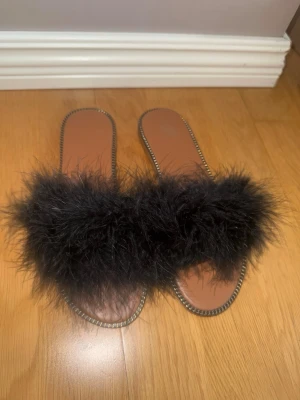 Svarta fluffiga tofflor med fjädrar - Säljer ett par svarta tofflor med fluffiga fjädrar över foten och dekorativ kant runt sulan. Sulan är brun med vågigt mönster undertill för bättre grepp. Perfekta för dig som vill ha något extra till hemmastilen. Helt nya aldrig använda.