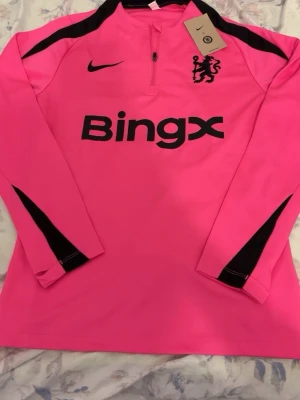 Rosa långärmad Nike träningströja - Snygg rosa långärmad träningströja från Nike med svarta detaljer och Bingx-logga på bröstet. Halvlång dragkedja framtill och svart lejonemblem på vänster sida. Perfekt för träning eller uppvärmning.