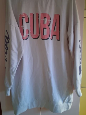 Vit hoodie med tryck CUBA - Vit hoodie med stort rosa och svart CUBA-tryck på ryggen och tryck på båda ärmarna. Lång modell med avslappnad passform och ribbade muddar. Perfekt för en chill och trendig look.