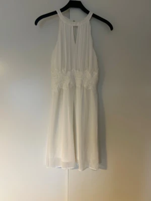 Vit ärmlös klänning med spetsdetaljer - Söt vit klänning med halterneck och vackra spetsdetaljer i midjan. Klänningen är ärmlös, har lätt vidd i kjolen och är tillverkad i ett tunt, flowy tyg som ger en luftig känsla. Perfekt för sommarens alla tillfällen. Men även perfekt som studentklänning. Använd en gång säljes på grund av att den inte används