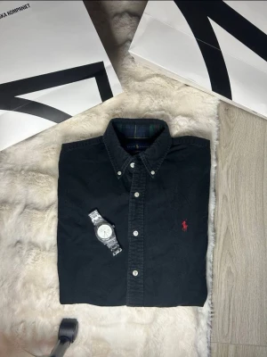 Svart skjorta från Polo Ralph Lauren - Klassisk svart skjorta från Polo Ralph Lauren med button-down krage och den ikoniska röda logotypen broderad på bröstet. Skjortan har vita knappar och är tillverkad i ett mjukt bomullstyg. Perfekt för en clean och stilren look. Slim fit storlek m skicka dm för fler bilder!