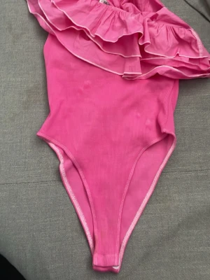 Rosa Zara body - Snygg rosa body med trendig volang upptill som ger en cool och lekfull look. Bodyn har en axelbandslös design och är tillverkad i ett stretchigt material som sitter bekvämt. 