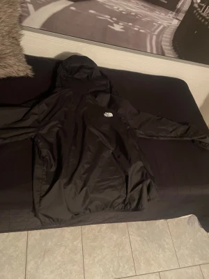 Svart vindjacka från The North Face - Snygg svart vindjacka från The North Face med huva och dragkedja. Jackan har en stilren design med logga på bröstet och är tillverkad i ett lätt, vindtätt material. Perfekt för blåsiga dagar och enkel att matcha med olika outfits.