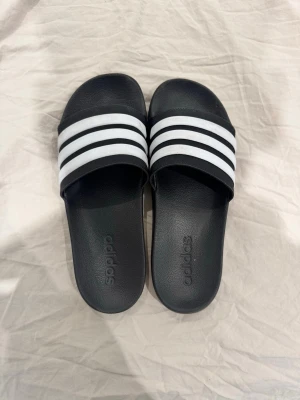 Svarta Adidas tofflor - Klassiska svarta Adidas badtofflor med tre vita ränder över foten och Adidas-logga på sidan. Tillverkade i syntetmaterial med räfflad sula för extra grepp. Perfekta för stranden, gymmet eller hemma. Litet hål som inte syns mycket. Använd kanske en sommar