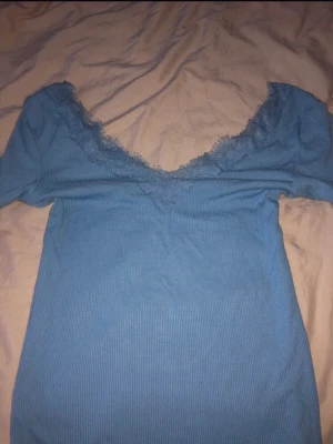 Blå ribbad topp med spetskant - Snygg blå ribbad topp med v-ringning och spetsdetaljer längs halsringningen. Toppen har korta ärmar och är perfekt för dig som gillar en feminin och trendig stil. Materialet känns mjukt och stretchigt.