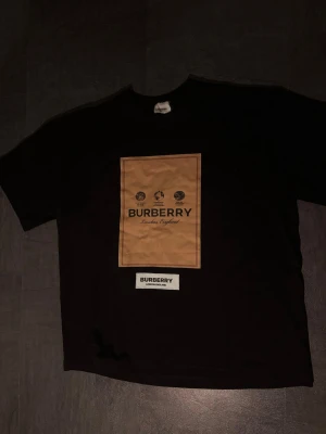 Svart Burberry t-shirt med patch - Snygg svart t-shirt från Burberry med stor beige patch på bröstet och svart text. Under patchen finns en vit etikett med Burberry-loggan. Klassisk rund hals och korta ärmar. Perfekt för dig som gillar exklusiva streetwear-plagg.