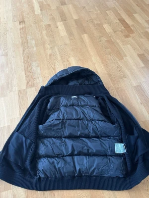 moncler cardigan herr sitter som L - Snygg svart pufferjacka med huva och stickade ärmar samt muddar. Jackan har quiltad framsida och baksida, dragkedja och ribbade detaljer. Perfekt för kalla vinterdagar och ger en sportig look.
