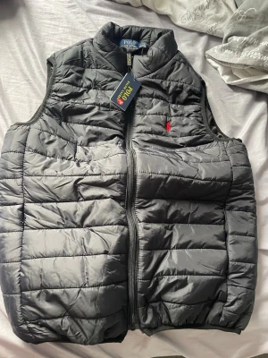 Svart dunväst från Polo Ralph Lauren - Snygg svart dunväst från Polo Ralph Lauren med klassisk quiltad design och hög krage. Västen har dragkedja framtill och den ikoniska röda loggan broderad på bröstet. Perfekt för lager-på-lager och streetstyle.OBS köpt helt ny för o sälja vidare. Xl fast passar också som L skulle jag säga 