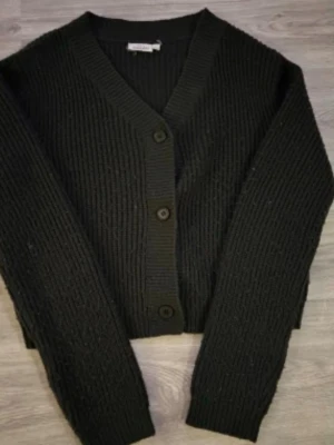 Svart stickad kofta från KappAhl - Mysig svart stickad kofta från KappAhl med v-ringning och tre knappar framtill. Koftan har ribbstickad struktur, långa ärmar och en något kortare passform. Perfekt att slänga över en t-shirt eller linne.