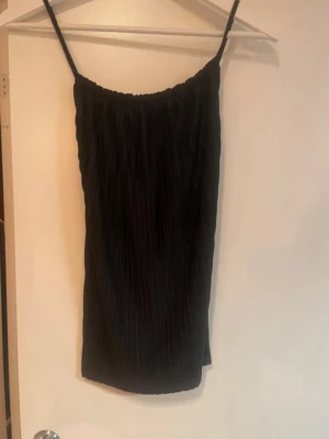 Svart plisserad linnetopp - Svart linnetopp med tunna axelband och plisserat tyg. Toppen är ärmlös och har en rak, loose passform som ger en avslappnad vibe. Perfekt att matcha med jeans eller kjol för en stilren look.