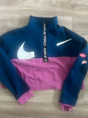 Nike Pro Dri-FIT träningströja M - Nike Pro Dri-FIT träningströja i blått och rosa med halv dragkedja och hög krage. Stora vita Swoosh-loggor på bröstet och ärmen, samt Nike Pro-tryck på dragkedjan. Tillverkad i funktionsmaterial som andas.