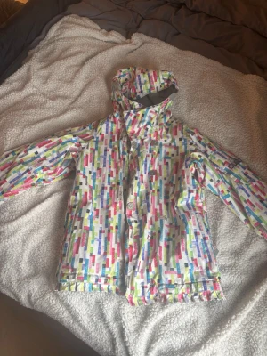 Färgglad vindjacka från Burton - Säljer en färgglad vindjacka från Burton med mönster i rosa, blått, grönt och gult. Jackan har huva, hög krage och dragkedja med knappar. Materialet är Dryride, perfekt för blåsiga dagar. Snygg och unik design som sticker ut.