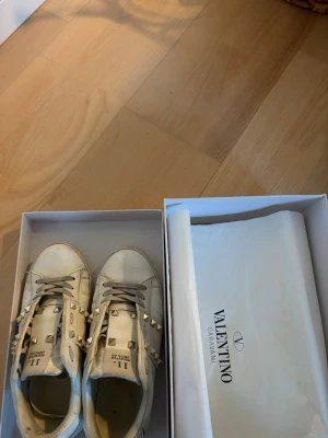 Vita Valentino sneakers med nitar - Snygga vita sneakers från Valentino Garavani med coola nitar längs sidorna. Skorna har klassisk låg modell, rund tå och vita snören. Tillverkade i läder och levereras i originalkartong. De saknas tyvärr 3 nitar och är lite slitna❤️❤️
