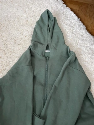 Grön hoodie med dragkedja Kappahl - Snygg grön hoodie från Kappahl med dragkedja framtill och stor huva. Tillverkad i mjukt sweatshirtmaterial och har en klassisk känguruficka. Perfekt för en chill och avslappnad stil.