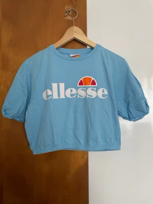 Blå croppad t-shirt från Ellesse - Snygg ljusblå croppad t-shirt från Ellesse med stort vitt tryck och logga framtill. T-shirten har korta ärmar och resår i nederkanten för en trendig passform. Perfekt för dig som gillar sportig och avslappnad stil.