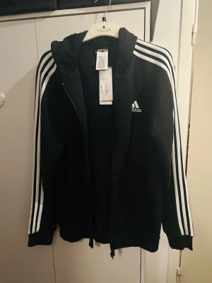 Svart Adidas hoodie med dragkedja - Svart hoodie från Adidas med hel dragkedja och klassiska vita ränder längs ärmarna. Adidas-logga på bröstet och två fickor framtill. Tillverkad i mjukt material och har en huva med snörning. Perfekt för en sportig och avslappnad stil.