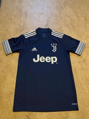 Juventus 2020/2021 Adidas - Juventus officiella matchtröja i mörkblått med vita detaljer och tre ränder på axlarna. Adidas-logga och Jeep-tryck på bröstet, samt Juventus klubbmärke med tre stjärnor. Tillverkad i lätt och ventilerande material.