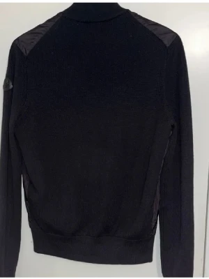 Svart cardiganjacka från moncler - Svart cardigan black label från moncler med stickade ärmar och rygg samt glansig framsida i syntetmaterial. Jackan har dragkedja framtill och två fickor med dragkedja. Klassisk logotyp på ärmen och ribbade muddar.