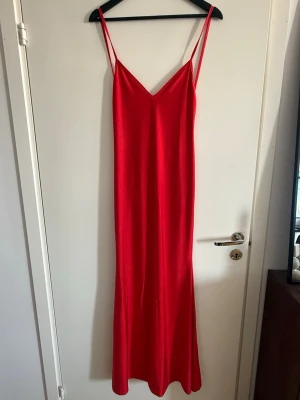 Röd lång klänning med smala axelband - Säljer en röd långklänning med smala axelband och v-ringning både fram och bak. Klänningen har en enkel, elegant design och är lätt utsvängd nertill. Materialet ser ut att vara mjukt och följsamt, perfekt för sommarens varma dagar.