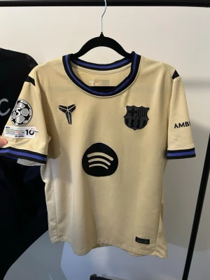 FC Barcelona bortatröja 2025/2026 - Säljer en riktigt snygg FC Barcelona bortatröja säsong 24/25 med Lamine Yamal #10 på ryggen. Beige/guldig färg med svarta och blå detaljer. Champions League patches på ärmen och Spotify sponsor framtill.