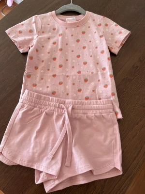 Rosa set med jordgubbsmönster - Sött rosa set med t-shirt och shorts. T-shirten har ett lekfullt jordgubbsmönster med små vita blommor och rund hals. Shortsen är enfärgade rosa med dragsko i midjan. Materialet är mjukt bomullstyg som känns bekvämt mot huden.