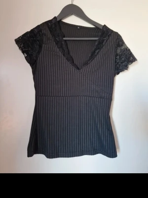 Svart topp med spetsdetaljer - Svart ribbad topp med v-ringning och spetsdetaljer vid ärmar och hals. Kortärmad modell med figurnära passform och omlottkänsla framtill. Perfekt för dig som gillar en stilren men ändå lite extra look. Storlek M men jag som vanligtvis har S-M i mindre toppar tyckte att det var för tajt. Några lösa trådar men på hela taget i väldigt fint skick. Katt finns i hemmet. Jag har fler toppar till salu