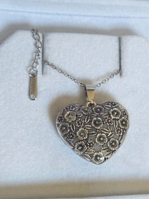 Silverfärgat hjärthalsband med blommor - Unikt handgjort halsband med ett stort hjärtformat hänge i silverfärg. Hänget är dekorerat med detaljerade blommor i relief och har en tunn kedja i rostfritt stål med justerbar längd. Hänget ger ifrån sig ett lila sken i mörker efter att ha varit i Dagsljus eller solljus en liten stund. Perfekt för dig som gillar romantiska och boho-inspirerade accessoarer.