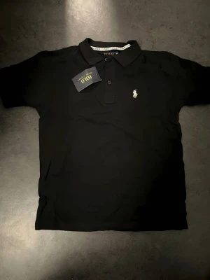 Svart pikétröja från Polo Ralph Lauren - Klassisk svart pikétröja från Polo Ralph Lauren med vit broderad logga på bröstet. Tröjan har krage, knappar framtill och korta ärmar. Perfekt till både jeans och shorts för en clean och stilren look.