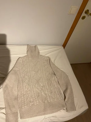 Beige kabelstickad polotröja - Säljer en beige kabelstickad polotröja med hög krage och ribbade muddar. Tröjan har ett klassiskt mönster framtill och är gjord i ett mjukt stickat material. Perfekt för att hålla sig varm under kyliga dagar.