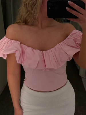 Rosa offshoulder volangtop - Säljer en ljusrosa offshoulder topp med volangdetalj över bröstet. Toppen är croppad och har en romantisk känsla med ärmlös design och bred volang. Perfekt för dig som vill ha en söt och trendig look.
