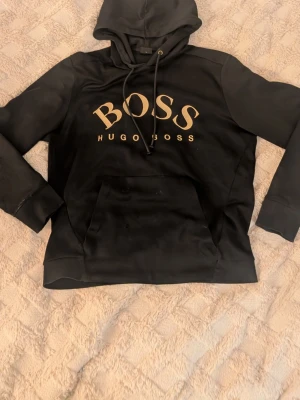 Svart Hugo Boss hoodie  - Snygg svart hoodie från Hugo Boss med gulddetaljer, lite slitage på armarna men inga hål 
