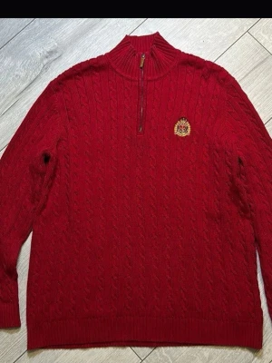 Röd kabelstickad tröja Ralph Lauren - Röd kabelstickad tröja från Ralph Lauren med halv dragkedja och broderad guldfärgad logotyp på bröstet. Tröjan har hög krage och ribbade muddar. Perfekt för dig som gillar klassisk och preppy stil. Pris kan diskuteras storlek L