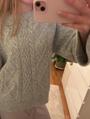 Ljusgrå kabelstickad tröja - Mysig ljusgrå stickad tröja med klassiskt kabelstickat mönster framtill. Inte använd så mycket. Tröjan har rund halsringning, långa ärmar och ribbade muddar vid ärmslut och nederkant. Perfekt för kyliga dagar och enkel att matcha med olika outfits.