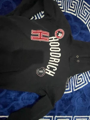 Svart hoodie från Hoodrich med tryck - Svart hoodie från Hoodrich med stora vita och röda tryck framtill, inklusive ett stort H och flera patchar. Klassisk huva med snörning och långa ärmar. Perfekt för en streetwear-look.